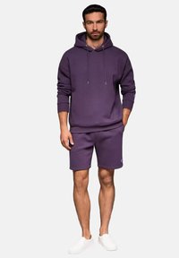 Lila Baumwollkapuzenpullover mit Fronttasche und Zugbändern, kombiniert mit passenden lila Shorts. Das Model trägt weiße Slipper.