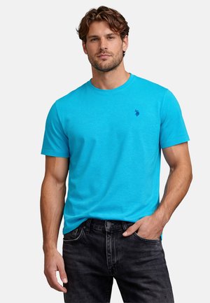 Man wearing bright blue short-sleeve crew neck t-shirt and black jeans, standing with one hand in pocket against plain background.