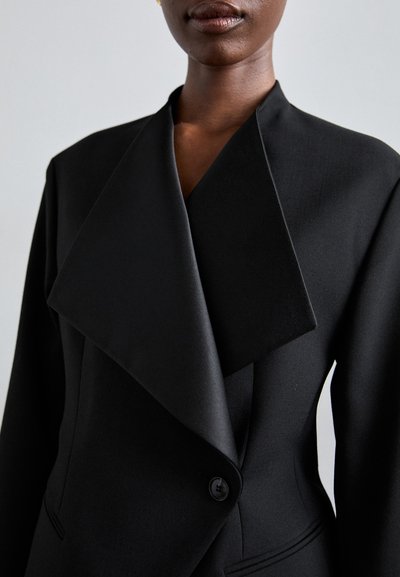 Μαύρο σακάκι που έχει φαρδιά γιακα, κλείσιμο με μονό κουμπί και λεία υφή υφάσματος. Περιλαμβάνει λεπτομέρεια πλάι τσέπης.