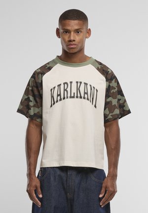 Homme portant un t-shirt beige avec le texte « KARL KANI » et des manches camouflage, associé à un jean bleu foncé, debout devant un fond gris.