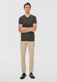 Czarna oliwkowa koszulka typu V-neck, beżowe spodnie slim-fit i czarne buty; model stoi z założonymi rękami, neutralne tło.