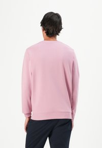 Vyras su tamsiais plaukais, apsirengęs šviesiai rožinės spalvos ilgos rankovės sweatshirt ir tamsiomis kelnėmis, stovi atsukęs nugarą prieš baltą foną.