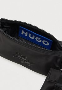 Μαύρη θήκη με φερμουάρ και ανάγλυφο "Hugo" στο μπροστινό μέρος, με μπλε εσωτερική ετικέτα που γράφει "HUGO" με ασημένια γράμματα.