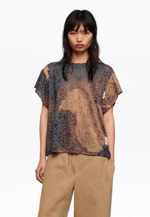 Joven con cabello negro y lacio que lleva una camiseta de malla de manga corta con estampado sobre pantalones beige, de pie frente a un fondo blanco liso.