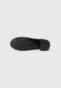 Chaussure à semelle en caoutchouc noir avec un dessus en cuir vert lisse et brillant, présentant un bout arrondi et des détails de couture minimalistes sur les côtés.
