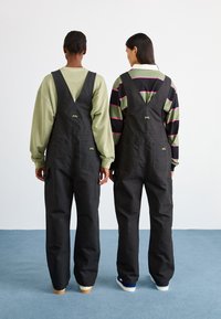 Zwarte katoenen tuinbroeken met een losse pasvorm, voorzien van verstelbare schouderbanden, twee voorkezen en geborduurde logo-accenten aan de achterkant.