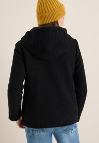 Veste en polaire noire avec capuche, présentant un tissu texturé, des manches longues et un ourlet arrondi. Portée avec un jean bleu et un bonnet jaune.