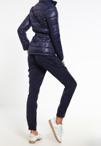 Granatowa kurtka puchowa o fakturowanym wykończeniu i elastycznym pasku w talii, zestawiona z granatowymi dresami i białymi sneakersami z jasnzielonymi akcentami.