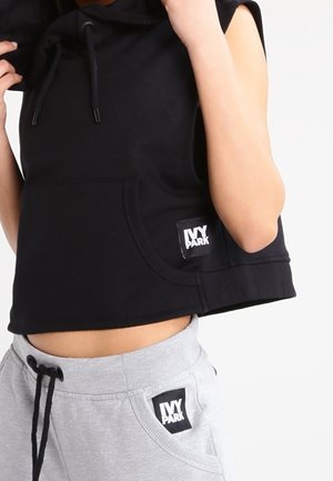 Osoba nosząca czarną bezrękawnikową bluzę z kapturem i szare spodnie dresowe, oba z logo Ivy Park na kieszeniach, pozująca z uniesionymi rękami.