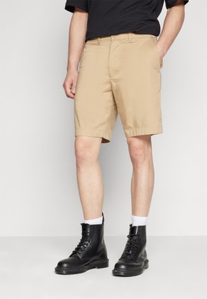 Person wearing beige knee-length shorts, black lace-up boots, white socks, and a black shirt, standing with hands in pockets.