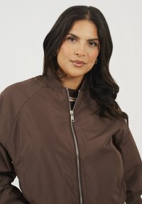 Brązowa kurtka bombowa wykonana z lekkiego materiału, z przodu zapinana na zamek, z prążkowanym kołnierzem i bocznymi kieszeniami, charakteryzująca się gładkim, matowym wykończeniem.