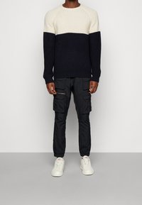 Pull en maille bleu marine et crème avec un motif texturé, associé à un pantalon cargo noir à multiples poches et des baskets blanches.