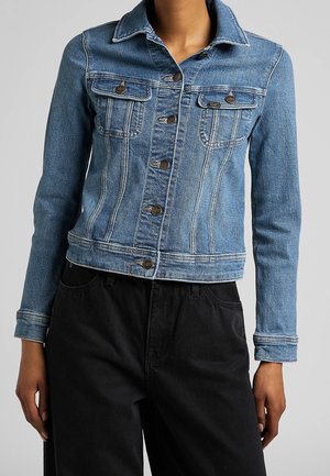 Personne portant une veste en jean bleue ajustée avec des boutons et des poches poitrine, associée à un pantalon noir taille haute, sur un fond uni.
