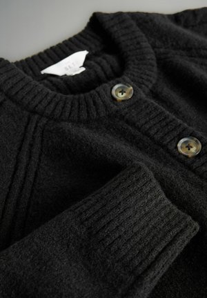 Schwarzer Wollcardigan mit einem gerippten Kragen, der vorne zwei große, runde, undurchsichtige Knöpfe aufweist. Weiche Textur und leicht lockere Passform.