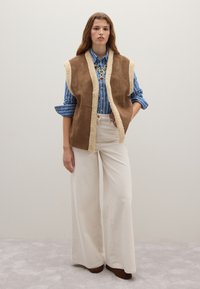 Brauner Wildlederweste mit Fellfutter, getragen über einem blauen gestreiften Button-Up-Hemd und cremefarbenen weitem Hosen. Das Model steht auf einem hellen Teppich.