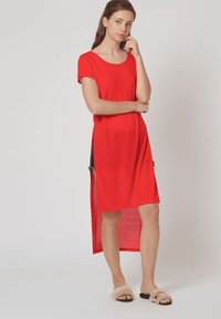 Vestido camisero rojo con cuello redondo, mangas cortas, aberturas laterales y una parte trasera ligeramente más larga. Lleva unas sandalias de diapositivas de felpa.