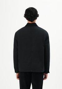 Man with short curly hair wears a black jacket and black pants, standing facing away against a plain white background.
