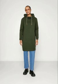 Vestito con cappuccio verde, dettagli con coulisse, maniche lunghe e tasche laterali, abbinato a jeans blue chiaro a gamba dritta e scarpe nere.