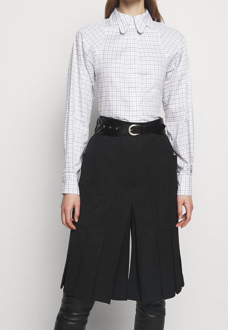 Camisa de cuadros blancos con cuello puntiagudo, combinada con una falda negra con pliegues. Presenta un cinturón ancho negro con una hebilla plateada, creando un aspecto estructurado.