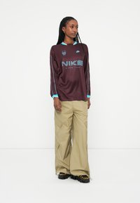 Dolga majica burgundske barve Nike z turkiznimi detajli in logotipom, v kombinaciji s svetlobeznimi širokimi hlačami. Črni čevlji z rumenimi vezalkami zaokrožijo videz.