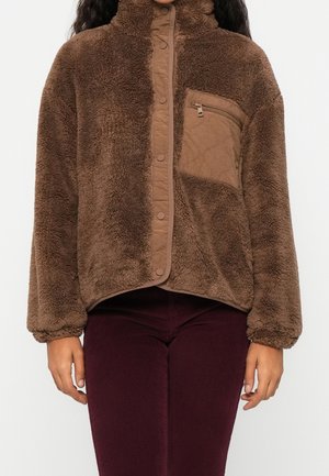 Person wearing a brown fuzzy zip-up jacket with a high collar and a quilted chest pocket, paired with dark maroon pants.