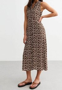 Ruskea hihaton mekko, jossa on valkoisia pilkkuja, napitettava etuosa ja A-linjainen siluetti, yhdistettynä ruskeisiin flip-flop -sandaleihin. Sileä kangas.