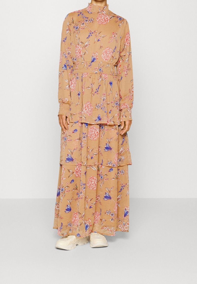 Femme porte une longue robe beige à fleurs roses et violettes, manches longues, col montant et baskets blanches, debout sur fond blanc.