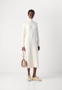 Robe en tricot blanche longue avec un col haut et des accents côtelés, associée à des baskets beige et un sac à main beige assorti. Texture lisse.