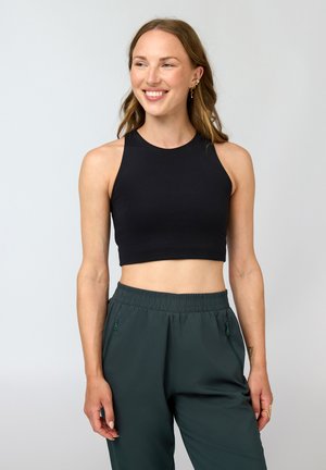 Smilende kvinne iført svart ermeløs crop top og mørkegrønne bukser med elastisk midje mot ensfarget lys bakgrunn.