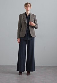 Brauner maßgeschneiderter Blazer, dunkel gestreiftes Hemd und weite Hose. Das Outfit weist klare Linien auf, mit einer glatten Textur und schichtweise gestaltet.