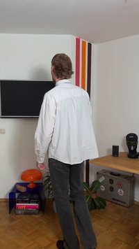 Chemise à rayures blanches, pantalon gris et chaussures marron. Lampe orange sur une table transparente, avec des livres en dessous. Mur orné de bandes de couleur vive.