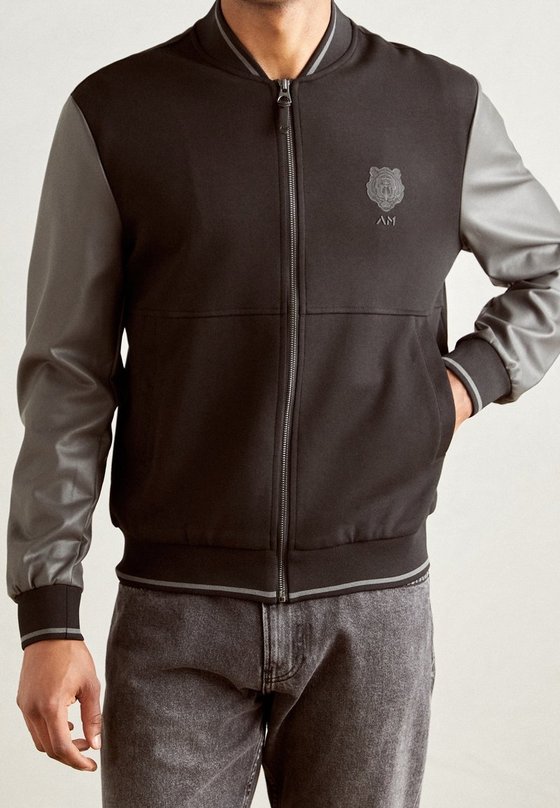 Homme portant une veste noire zippée avec des manches en cuir grises et des poignets et ourlet bordés de gris, associée à un jean gris.