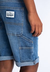 Leren spijker shorts in lichtblauw met opgerolde boorden, voorzien van meerdere zakken en oranje stiksels. Merkenlabel zichtbaar op de achterzak.