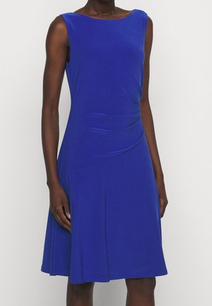 Vrouw draagt een mouwloze, knie-lange koningsblauwe jurk met een geplooid detail aan de rechterkant ter hoogte van de taille.