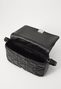 Sac à bandoulière en cuir matelassé noir avec rabat ouvert, montrant l'intérieur vide et la bandoulière ajustable sur fond blanc.