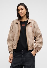 Ανοιχτό καφέ bomber μπουφάν με υφή, ελαστικά μανσέτα και μπροστινό φερμουάρ. Φορεμένο πάνω από μαύρο T-shirt με κόκκινο λογότυπο.