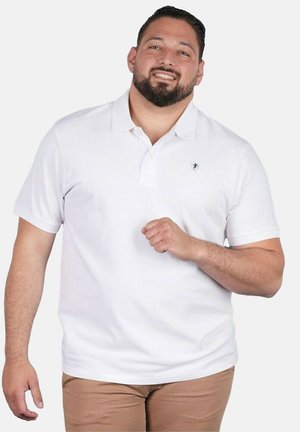 Homme souriant aux cheveux foncés et à la barbe, portant un polo blanc à manches courtes et un pantalon beige, debout devant un fond uni.