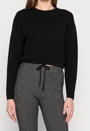 Femme portant un haut noir à manches longues et un pantalon en maille gris taille haute avec un cordon noir noué à la taille.