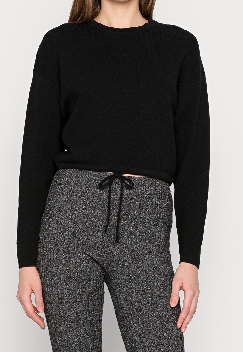 Femme portant un haut noir à manches longues et un pantalon en maille gris taille haute avec un cordon noir noué à la taille.