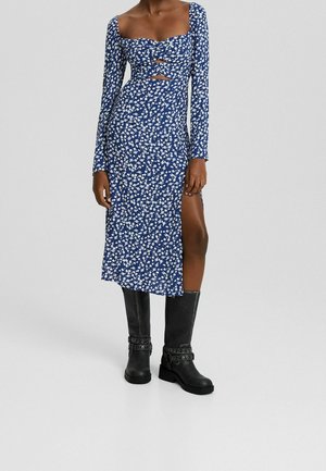 Femme portant une robe midi bleue à fleurs avec manches longues, découpes à l'avant, fente haute sur la cuisse, et bottes noires montantes à boucles.