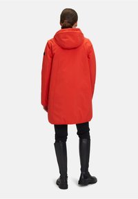 Manteau imperméable rouge avec capuche, d'une coupe ample et d'un design mi-long, associé à un pantalon noir ajusté et des bottes noires jusqu'aux genoux.