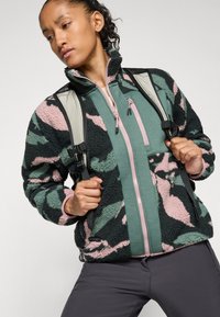Donna che indossa una giacca in pile camouflage verde e rosa, pantaloni grigi e uno zaino grigio, mentre tiene le cinghie dello zaino, guardando di lato.