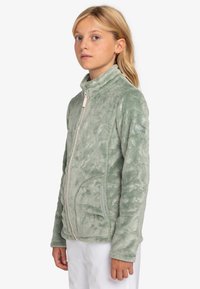 Chaqueta de forro polar verde claro con cuello alto, cremallera frontal y bolsillos laterales. Presenta un sutil patrón texturizado y el logo de Roxy en la manga.
