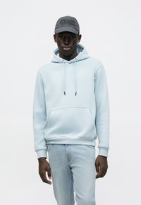 Lyseblå hettegenser med kengurulomme, justerbare snorer og ribbestrikkede mansjetter, kombinert med lyse denimbukser og en mørkegrå caps.