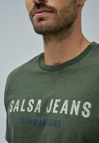 Camiseta de algodón color verde oliva con el texto "SALSA JEANS" en blanco y "DENIM ARTISANS" en azul, cuello redondo, textura suave.
