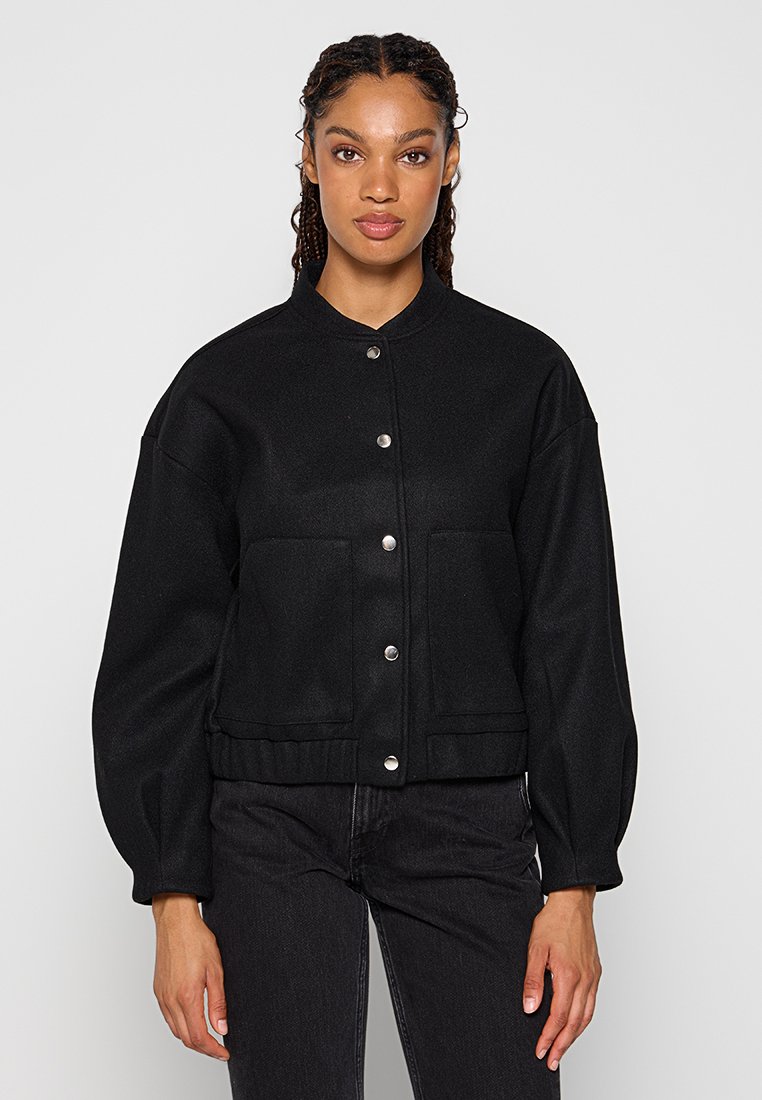 Schwarze verkürzte Jacke mit rundem Hals, silbernen Druckknöpfen und zwei Fronttaschen. Hergestellt aus strukturiertem Stoff mit lockerer Passform.