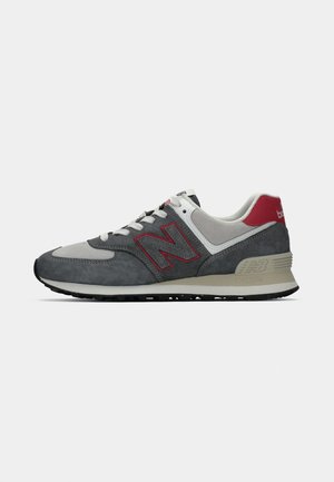 Zapatillas de ante gris con acentos rojos y una suela blanca. Presentan un collar acolchado, un diseño de cordones y un prominente logo "N" en el lateral.