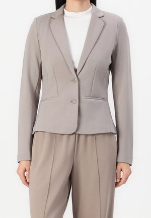 Kvinde iført en lysebeige skræddersyet blazer med to knapper over en hvid højhalset top og matchende beige plisserede bukser mod ensfarvet baggrund.