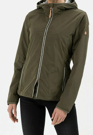 Personne portant une veste à capuche vert olive avec des fermetures éclair sur le devant et les côtés, associée à un pantalon noir, montrée des épaules aux hanches.