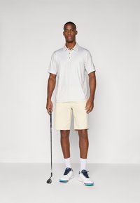 Hellgraues Polohemd mit kurzen Ärmeln und Knöpfen, beigefarbene Shorts und weiße Golfschuhe mit blauen Akzenten, gehalten von einer männlichen Figur mit einem Golfschläger.
